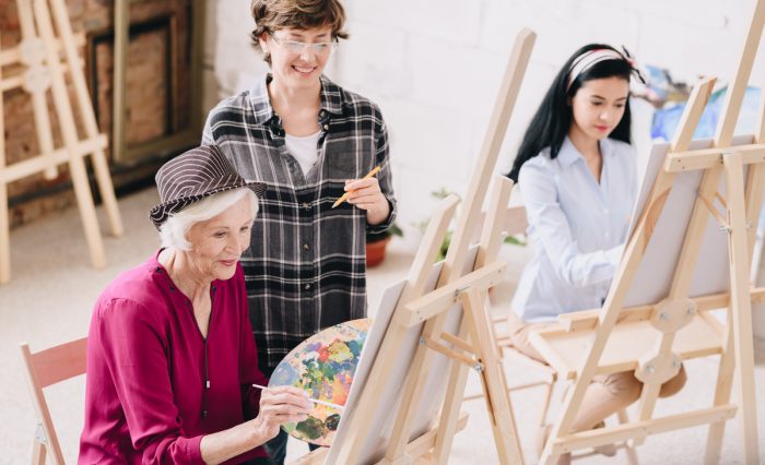Portrait of elegant senior woman painting sitting at easel in art studio with smiling female teacher giving comments, copy space (zh translation)